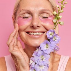 Vertical shot of fair haired mature lady smiles broadly shows white teeth touches face gently enjoys softness of skin applies hydrogel patches under eyes holds flower undergoes beauty procedures